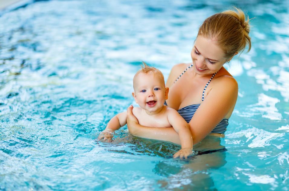 Mutter h&auml;lt l&auml;chelnd ein Baby im Wasser eines Schwimmbads, sanfte Wellen reflektieren das Licht.
