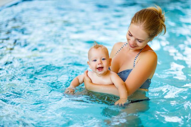 Mutter hält lächelnd ein Baby im Wasser eines Schwimmbads, sanfte Wellen reflektieren das Licht.