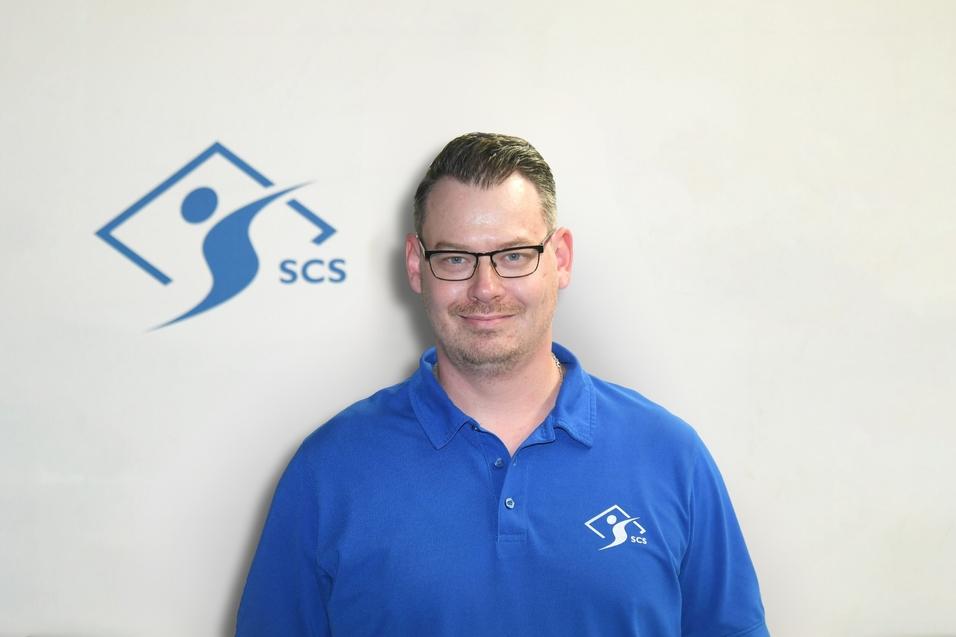 Mann mit Brille in blauem Polo-Shirt vor einer hellen Wand mit dem SCS-Logo.