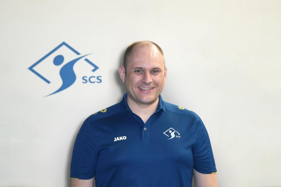 Mann in einem blauen Shirt mit Logo steht vor einer hellen Wand mit dem SCS-Logo. Er lächelt freundlich in die Kamera.