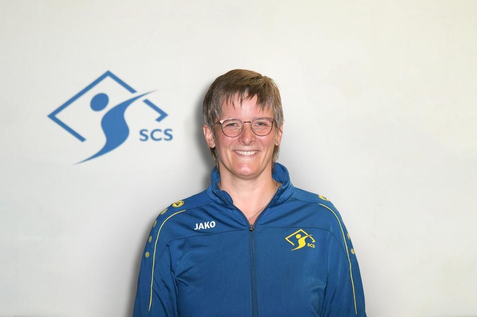 Portrait einer Person in einer blauen Trainingsjacke mit gelben Akzenten vor einem hellen Hintergrund mit dem Logo „SCS“.