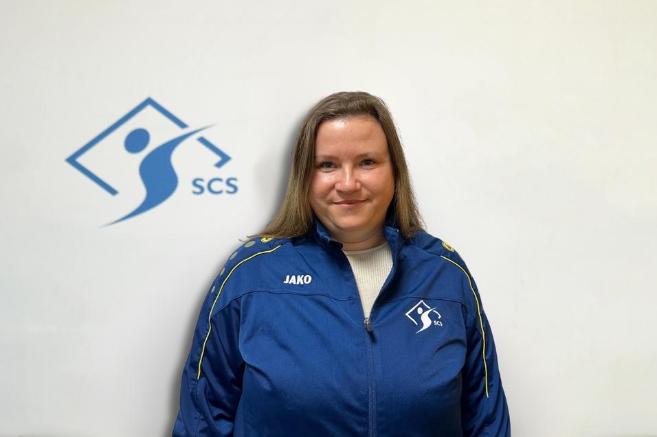 Porträt einer Frau in einer blauen Trainingsjacke mit Logo, vor einem hellen Hintergrund mit einem Symbol und der Aufschrift "SCS".