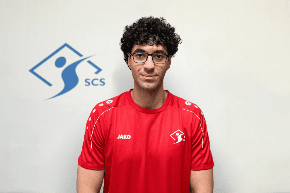 Porträt eines Mannes mit Locken, im roten Sporttrikot mit Logo, vor einer neutralen Wand mit SCS-Logo.