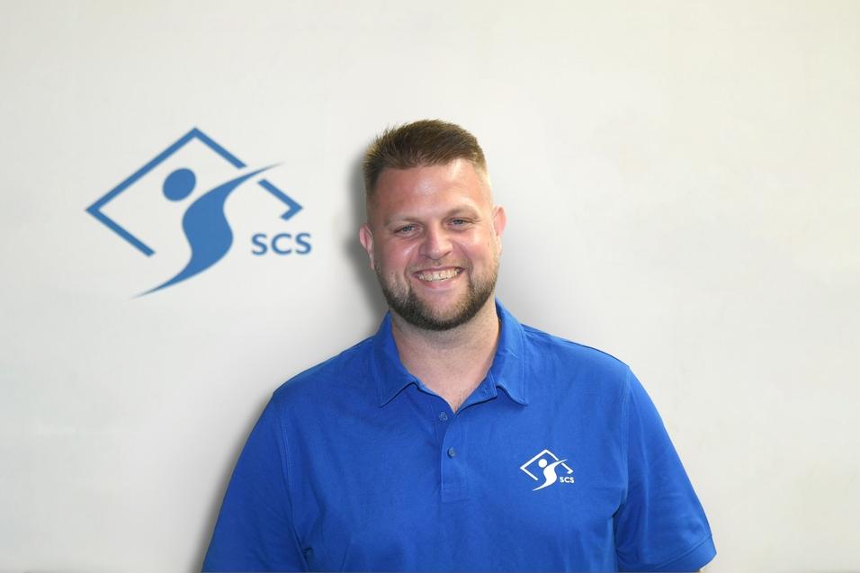 Lächelnder Mann in blauer Polo-Shirt vor hellgrauem Hintergrund mit SCS-Logo.