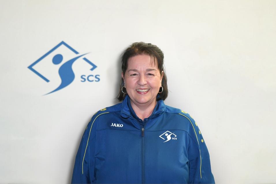 Frau in blauer Sportjacke mit Logo vor einem hellen Hintergrund, lächelt in die Kamera.