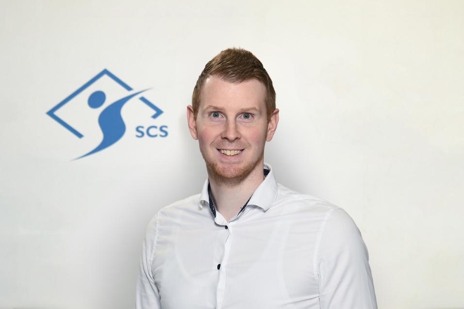 Porträt eines Mannes mit blondem Haar und weißem Hemd vor einem Hintergrund mit dem Logo „SCS“.