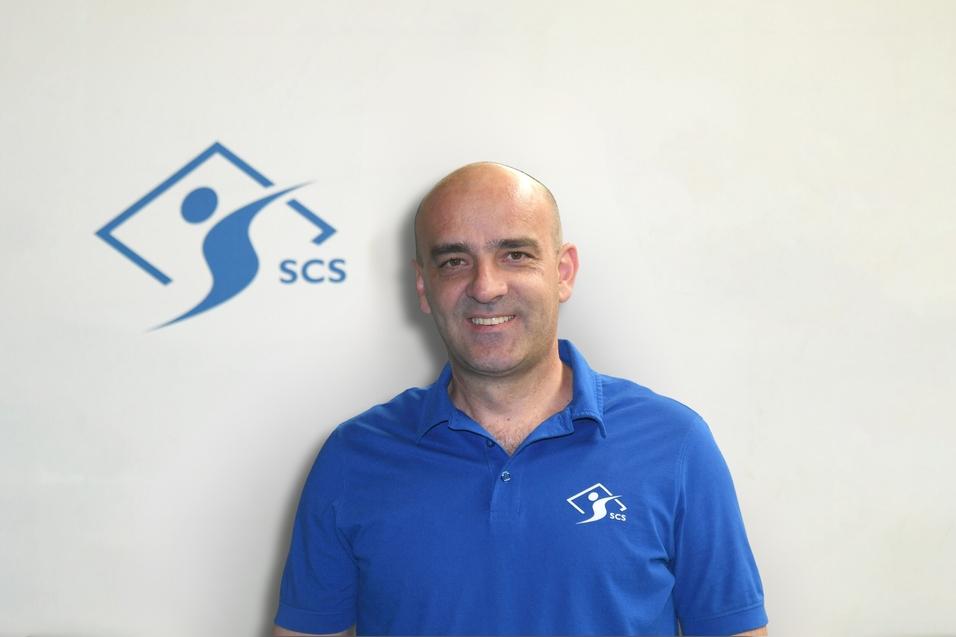 Mann mit Glatze trägt ein blaues Polo-Shirt mit Logo vor einer hellen Wand.