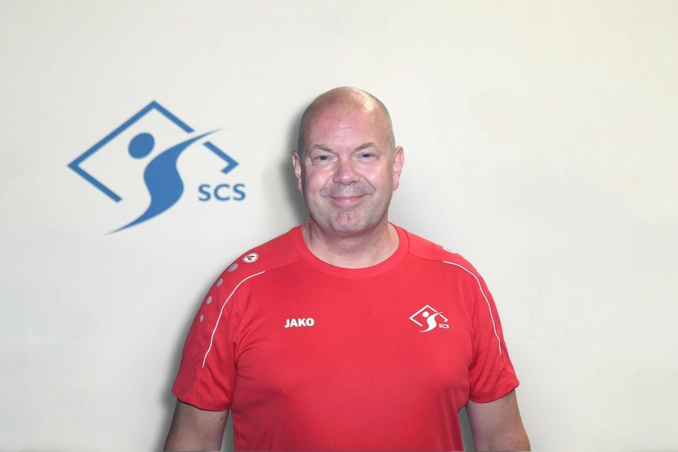 Mann mit Glatze in rotem Sportshirt steht vor einer hellen Wand mit einem SCS-Logo.