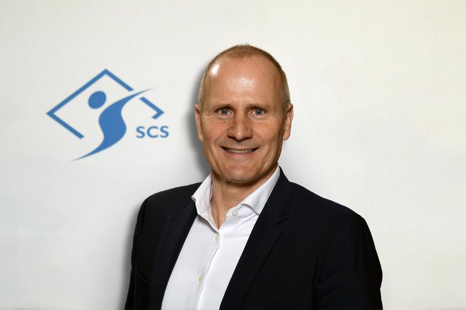 Mann in Blazer und Hemd lächelt vor hellem Hintergrund mit dem Logo der SCS.