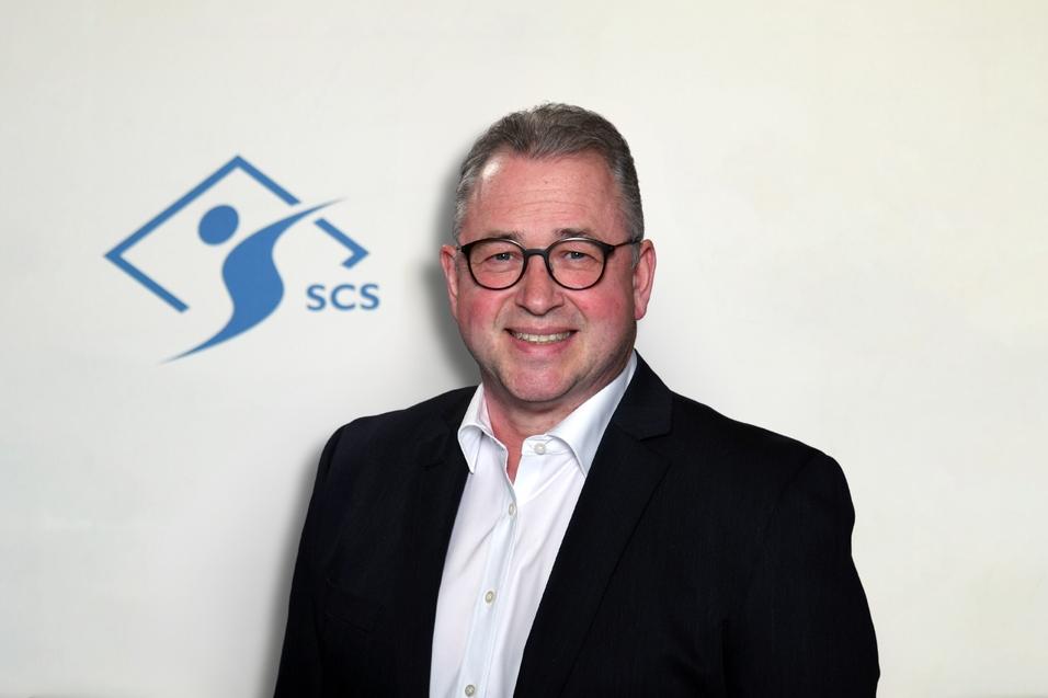 Porträt eines Mannes mit Brille in dunklem Anzug vor einem hellen Hintergrund mit dem SCS-Logo.