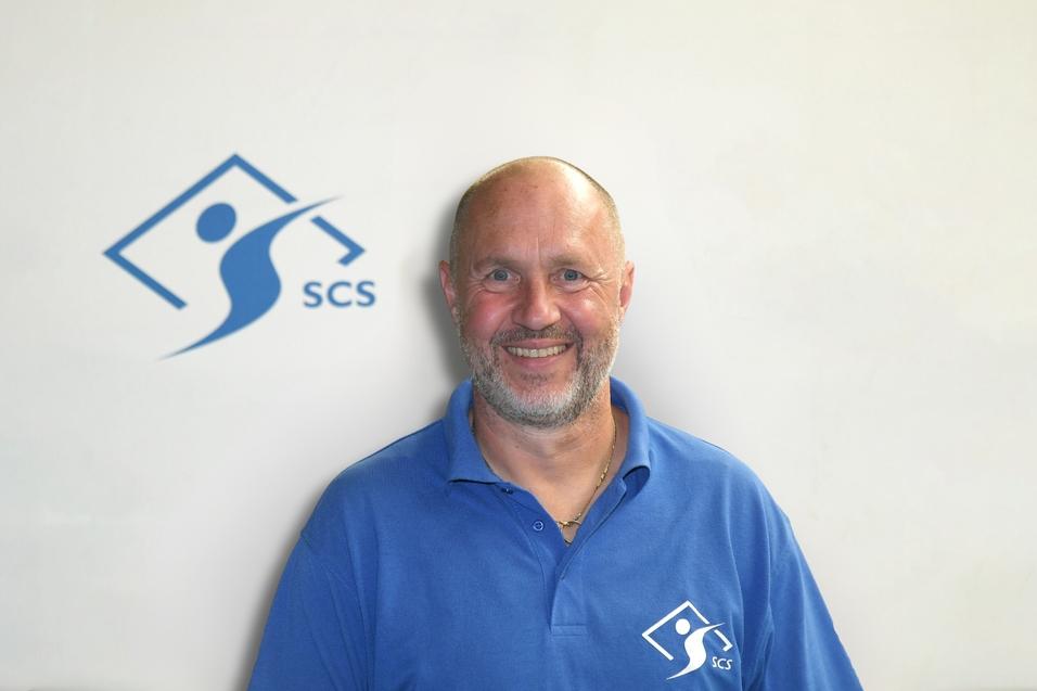 Mann mit kurzem Bart lächelt vor einem hellen Hintergrund, trägt ein blaues Hemd mit SCS-Logo.