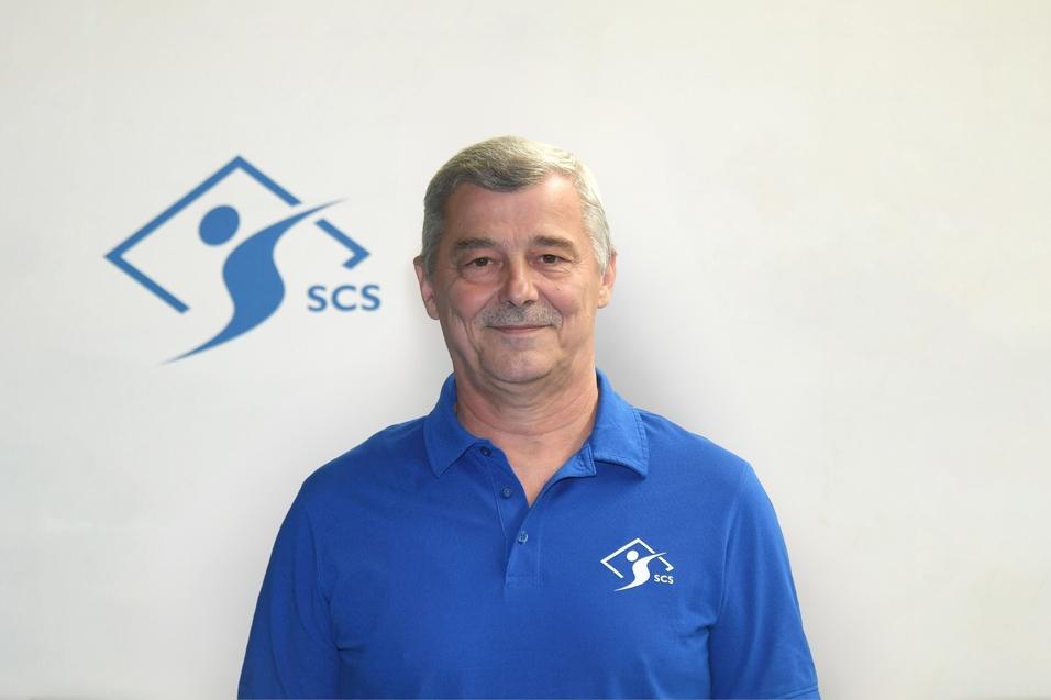 Mann in blauem Poloshirt mit Logo steht vor neutralem Hintergrund.