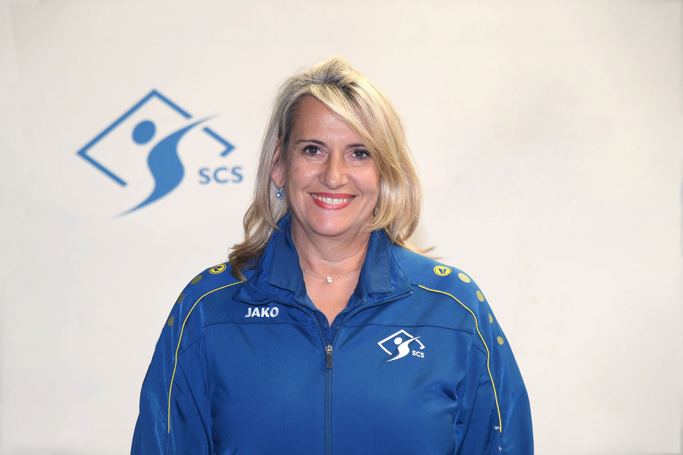 Frau mit blonden Haaren l&auml;chelt in blauer Sportjacke vor einem hellen Hintergrund mit SCS-Logo.
