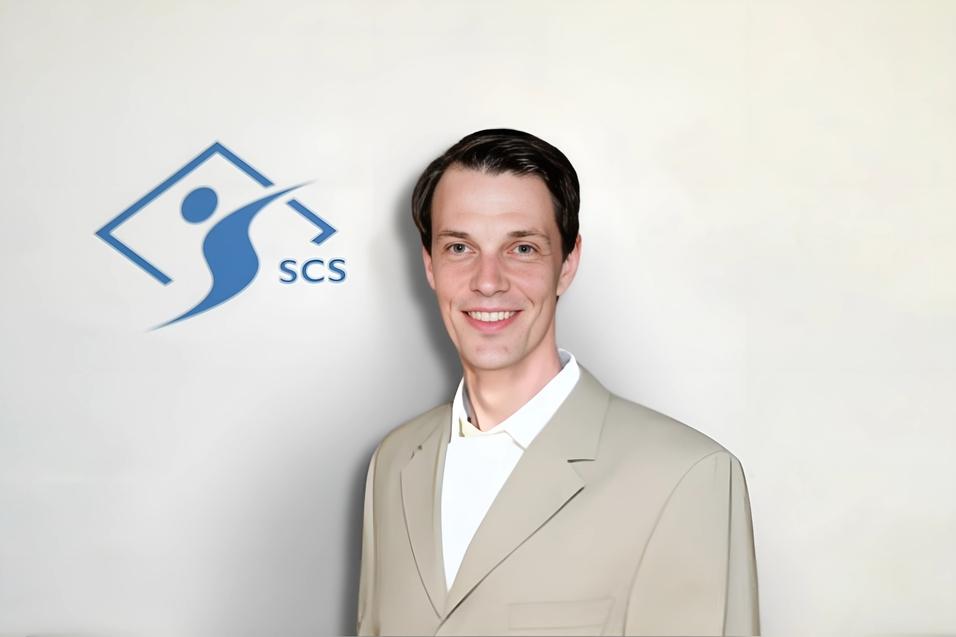 Mann im beigen Anzug lächelt vor einem hellen Hintergrund mit dem Logo „SCS“ links neben ihm.