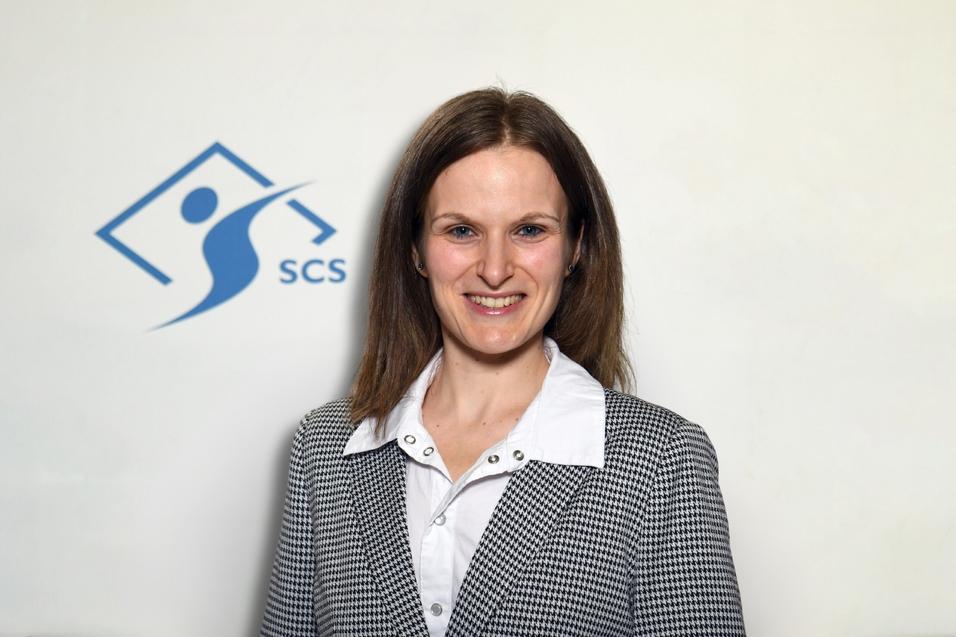 Portrait einer Frau mit langen Haaren, in einem karierten Blazer, vor einem Logo mit der Aufschrift "SCS".