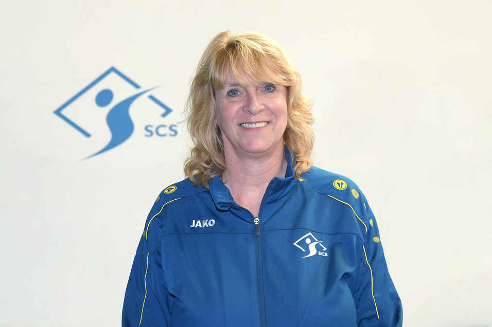 L&auml;chelnde Frau in blauer Jacke mit Logo, vor einem hellen Hintergrund mit SCS-Logo.