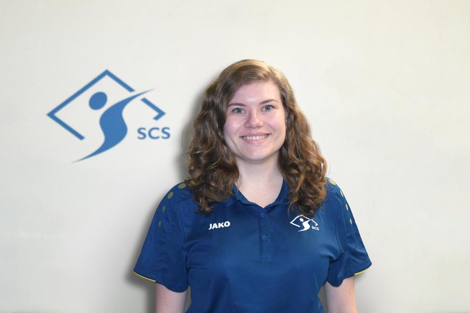 Junge Frau mit lockigem Haar trägt ein blaues Sportshirt mit Logo vor hellem Hintergrund.