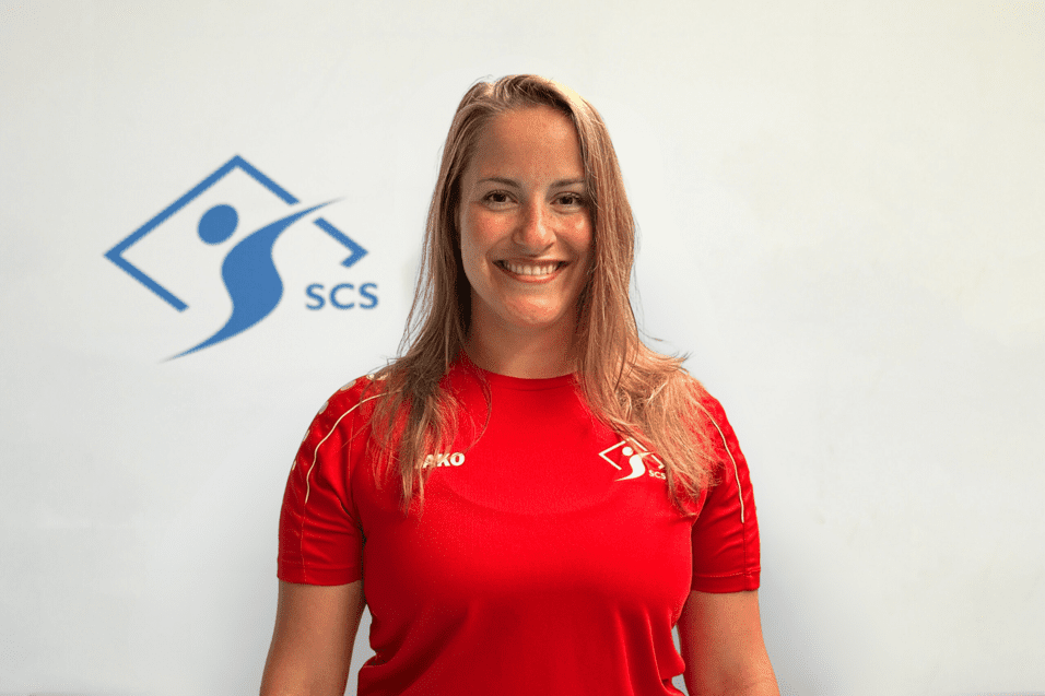 Portrait einer lächelnden Frau in rotem Sportshirt vor einem Logo mit der Aufschrift "SCS" auf hellem Hintergrund.