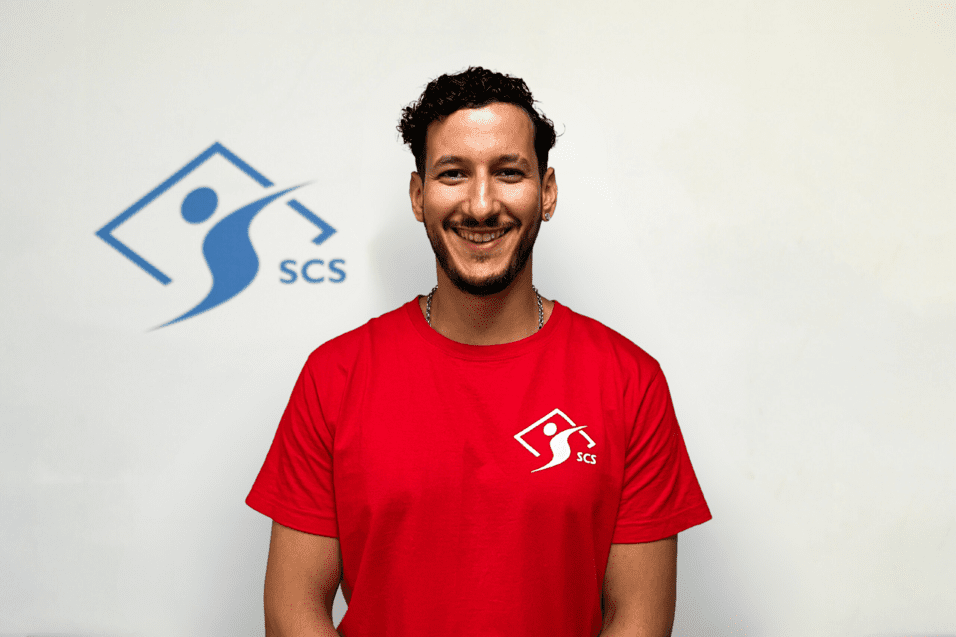 Lächelnder Mann in rotem T-Shirt mit SCS-Logo vor hellem Hintergrund.