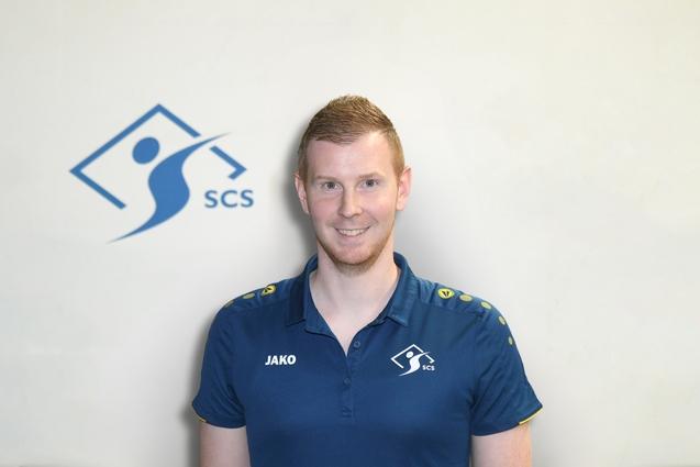 Porträt eines Mannes in einem blauen Poloshirt mit dem Logo „SCS“ vor einem hellen Hintergrund.