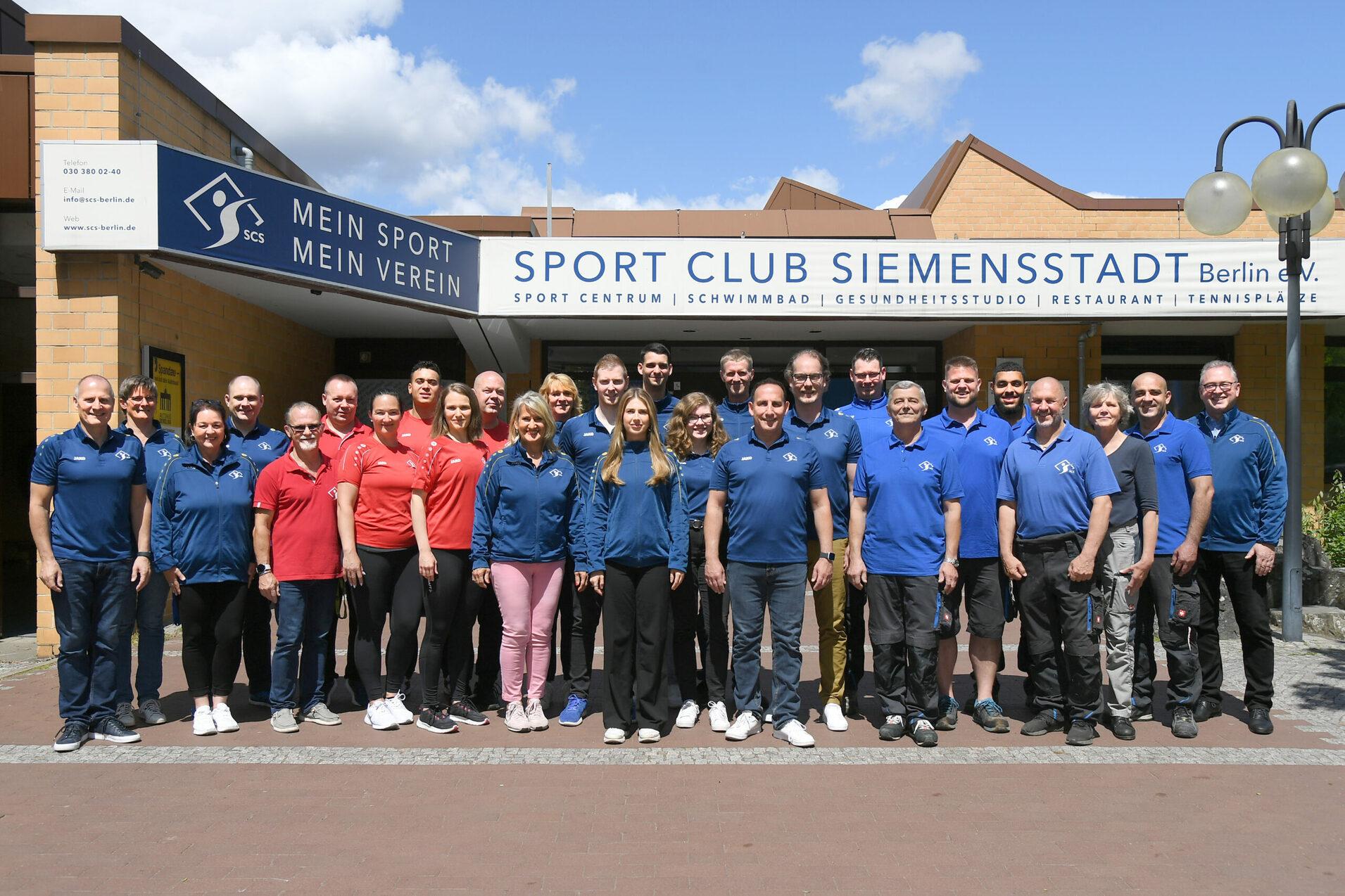Gruppenfoto von Mitgliedern des Sport Club Siemensstadt vor dem Vereinsgebäude, einige tragen rote und blaue Sportkleidung.