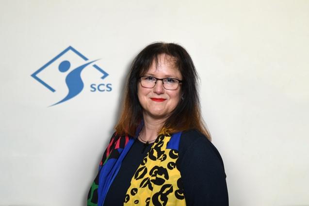 Porträt einer Frau mit Brille, farbenfrohem Schal und dunkler Kleidung vor einem SCS-Logo auf hellem Hintergrund.