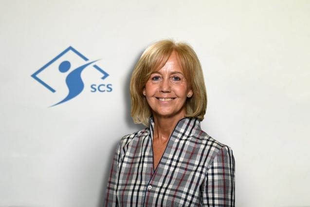 Porträt einer lächelnden Frau in kariertem Hemd, vor einem weißen Hintergrund mit SCS-Logo.