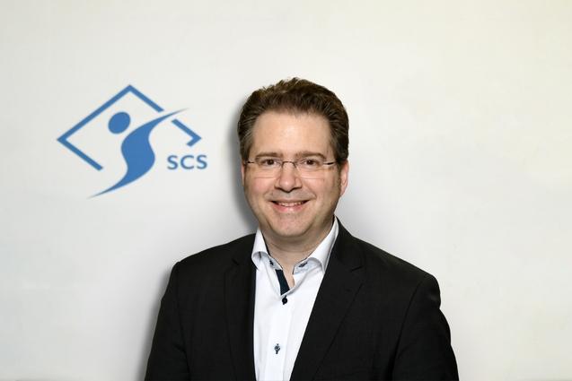 Porträt eines Mannes in Anzug, lächelnd vor einem hellen Hintergrund mit dem SCS-Logo.