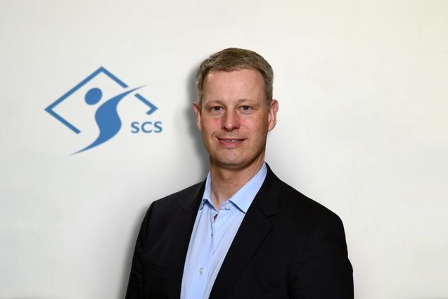 Porträt eines Mannes im Anzug vor einer hellen Wand mit dem Logo „SCS“ in Blau.