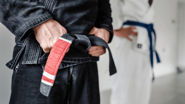 Nahaufnahme eines Sportlers mit schwarzem Gi, der seine rot-weiße Gürteltrage sieht, im Hintergrund ein weiterer Kämpfer.