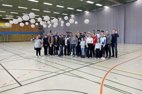 Gruppe von 25 Personen in einer Sporthalle, umgeben von schwebenden Laternen, alle l&auml;cheln und zeigen Daumen hoch.