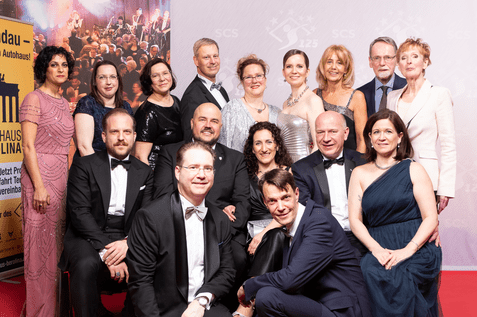 Gruppenaufnahme von 15 Personen in formeller Abendkleidung vor einem festlichen Hintergrund.