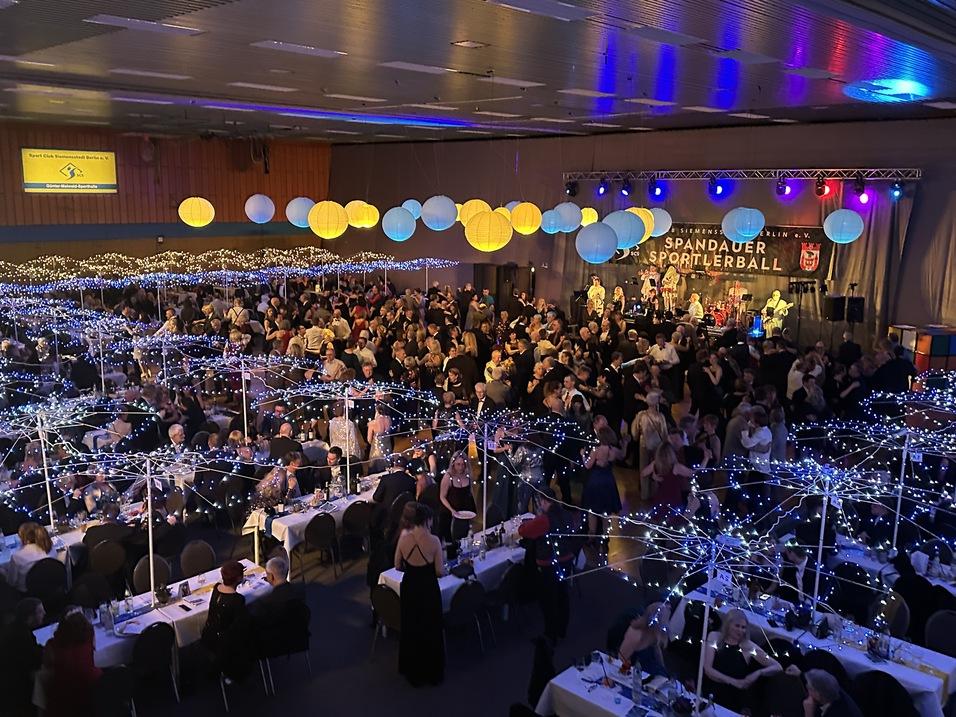 Große Veranstaltungshalle mit festlich geschmückten Tischen, Tänzern und einer Band auf der Bühne beim Spandauer Sportlerball.