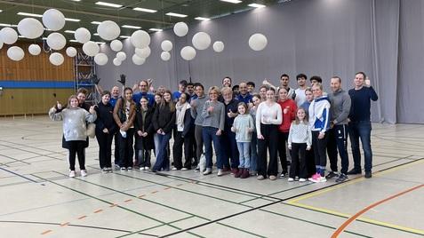 Gruppe von 25 Personen in einer Sporthalle, umgeben von schwebenden Laternen, alle lächeln und zeigen Daumen hoch.