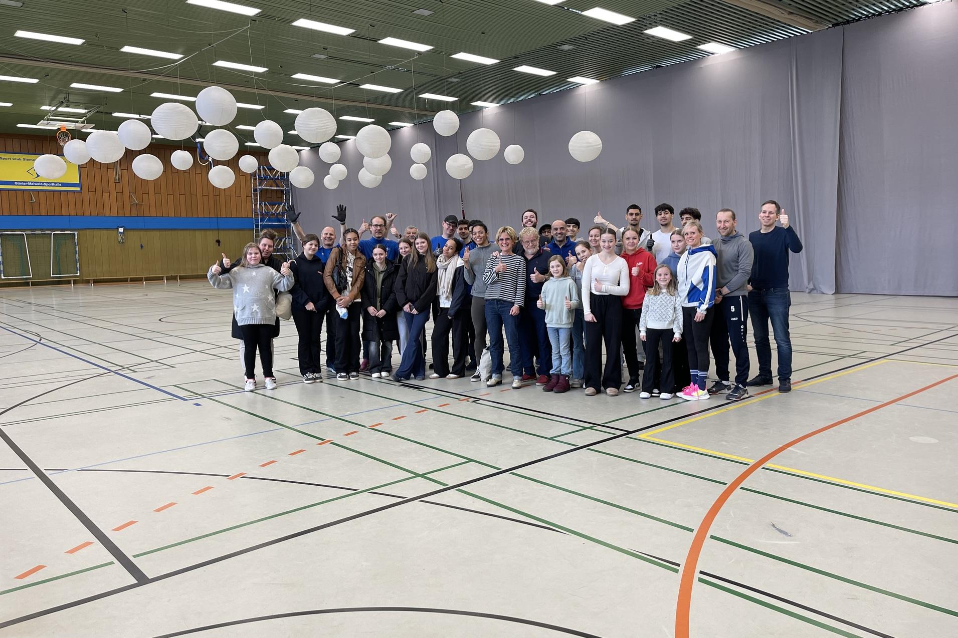 Gruppe von 25 Personen in einer Sporthalle, umgeben von schwebenden Laternen, alle l&auml;cheln und zeigen Daumen hoch.