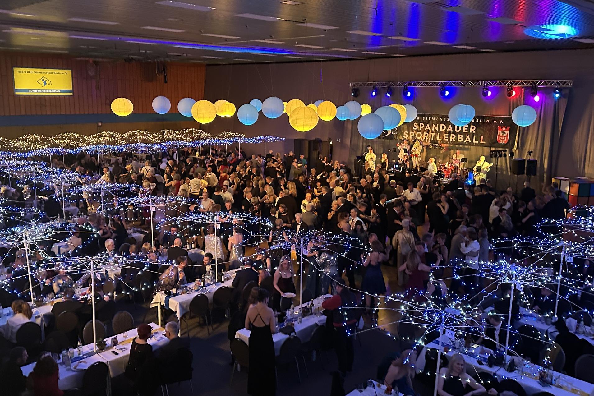 Gro&szlig;e Veranstaltungshalle mit festlich geschm&uuml;ckten Tischen, T&auml;nzern und einer Band auf der B&uuml;hne beim Spandauer Sportlerball.
