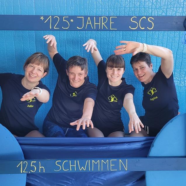 Vier Frauen in Sportshirts posieren fröhlich vor einem blauen Hintergrund mit der Aufschrift „125 Jahre SCS“ und „125 h Schwimmen“.