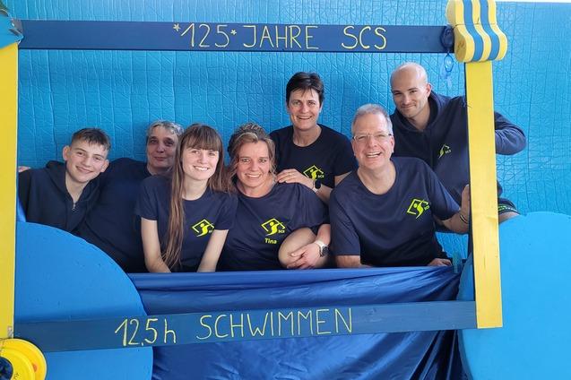 Gruppe von sechs Personen in einheitlichen T-Shirts, feiernd hinter einem Rahmen mit der Aufschrift „125 Jahre SCS“.