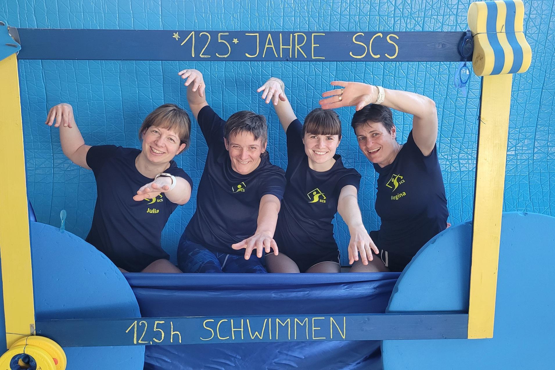 Vier Frauen in Sportshirts posieren fröhlich vor einem blauen Hintergrund mit der Aufschrift „125 Jahre SCS“ und „125 h Schwimmen“.