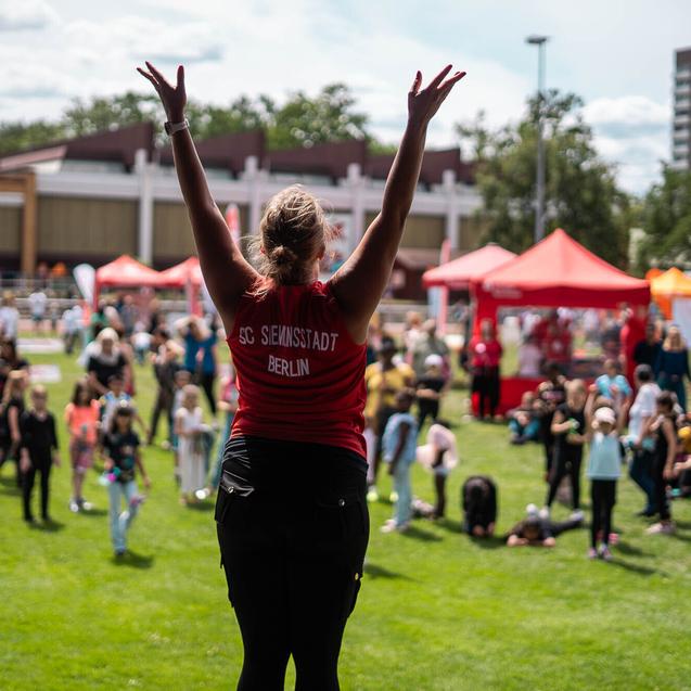 Eine Person in rotem Shirt hebt die Arme auf einer Festwiese, umgeben von einer Zuschauermenge und bunten Buden.