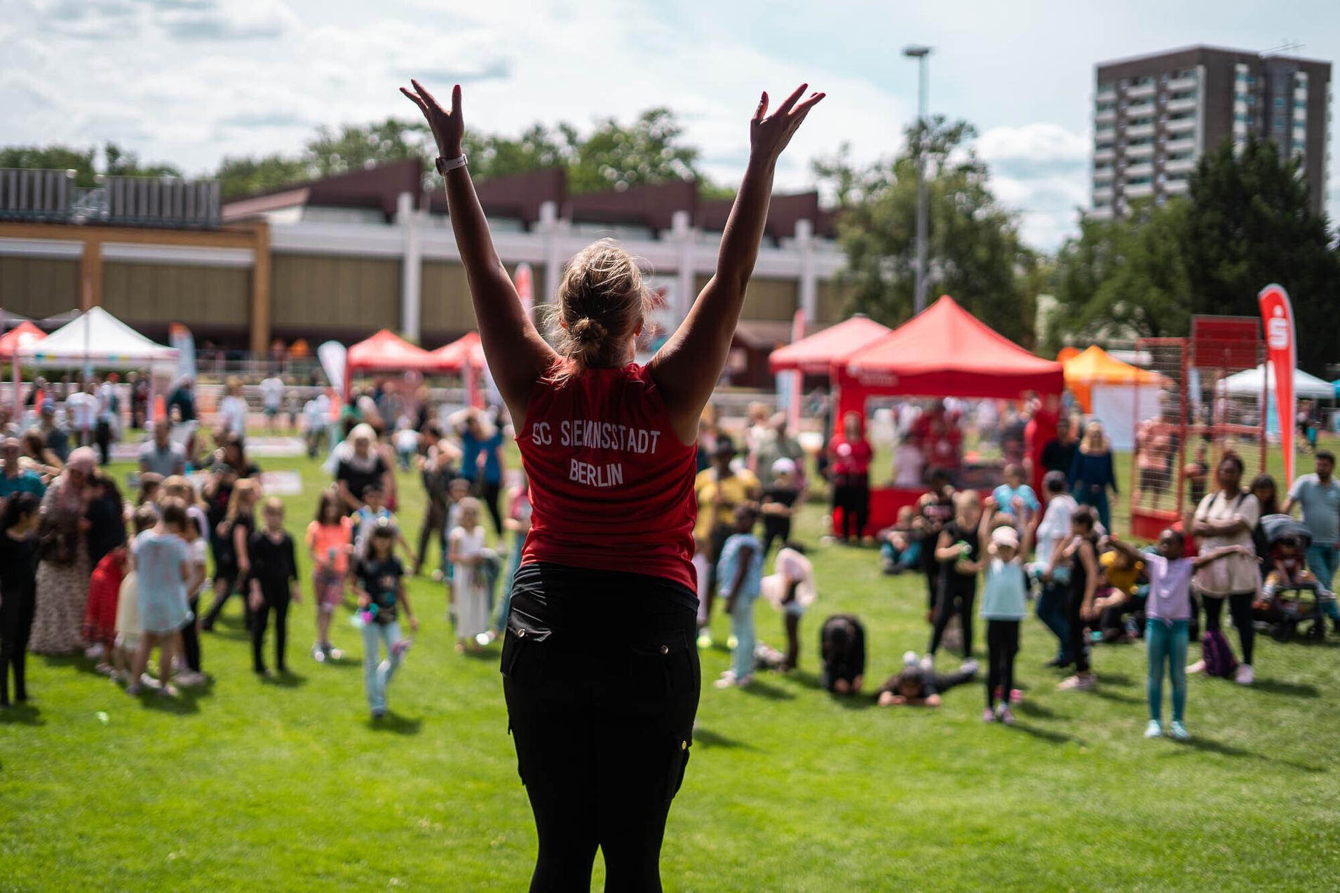 Eine Person in rotem Shirt hebt die Arme auf einer Festwiese, umgeben von einer Zuschauermenge und bunten Buden.