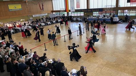 Tanzpaare in festlicher Kleidung auf einer Tanzfläche bei einem Wettbewerb, umgeben von Zuschauern und Tischen.