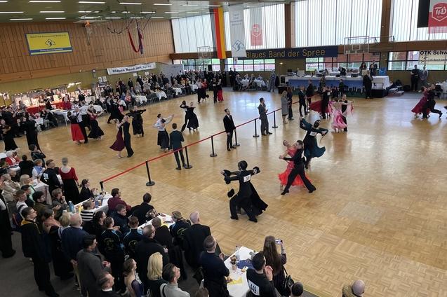 Tanzpaare in festlicher Kleidung auf einer Tanzfläche bei einem Wettbewerb, umgeben von Zuschauern und Tischen.