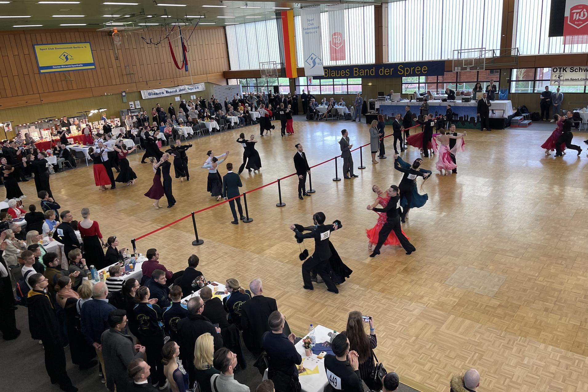 Tanzpaare in festlicher Kleidung auf einer Tanzfl&auml;che bei einem Wettbewerb, umgeben von Zuschauern und Tischen.