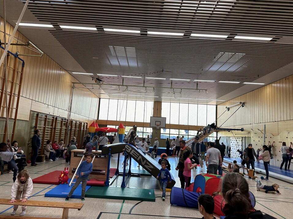 Viele Kinder und Erwachsene spielen in einer Sporthalle mit verschiedenen Geräten und Spielstrukturen.