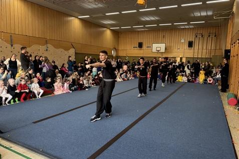 B&uuml;hnenauff&uuml;hrung in einer Turnhalle mit Zuschauern, die von Kindern in Kost&uuml;men umgeben ist.