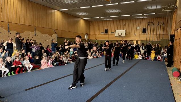 Bühnenaufführung in einer Turnhalle mit Zuschauern, die von Kindern in Kostümen umgeben ist.