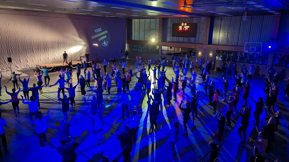 Gruppentanzveranstaltung in einer Sporthalle mit Teilnehmern unter bunten Lichtern und einem DJ auf einer Bühne.