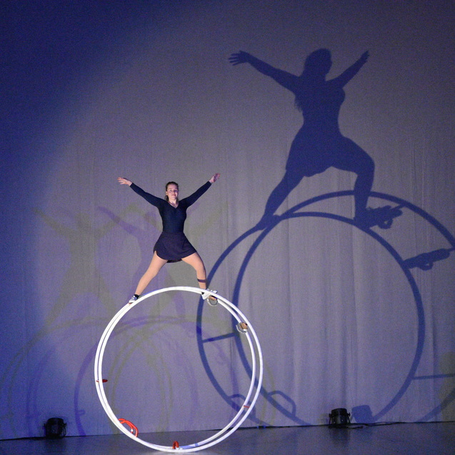 Akrobatin balanciert auf einer gro&szlig;en wei&szlig;en Requisite, w&auml;hrend Schatten an der Wand sichtbar sind.