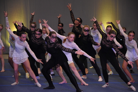 Gruppe junger Tänzerinnen in schwarz-weißen Kostümen mit Schminke, die synchron eine Tanzpose auf einer Bühne einnehmen.